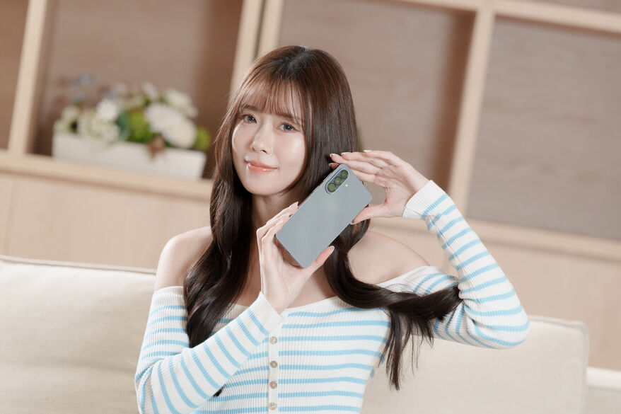 甜美模特兒身穿條紋露肩裝,雙手拿著 Galaxy A57 5G,展示其三鏡頭,笑容親切。